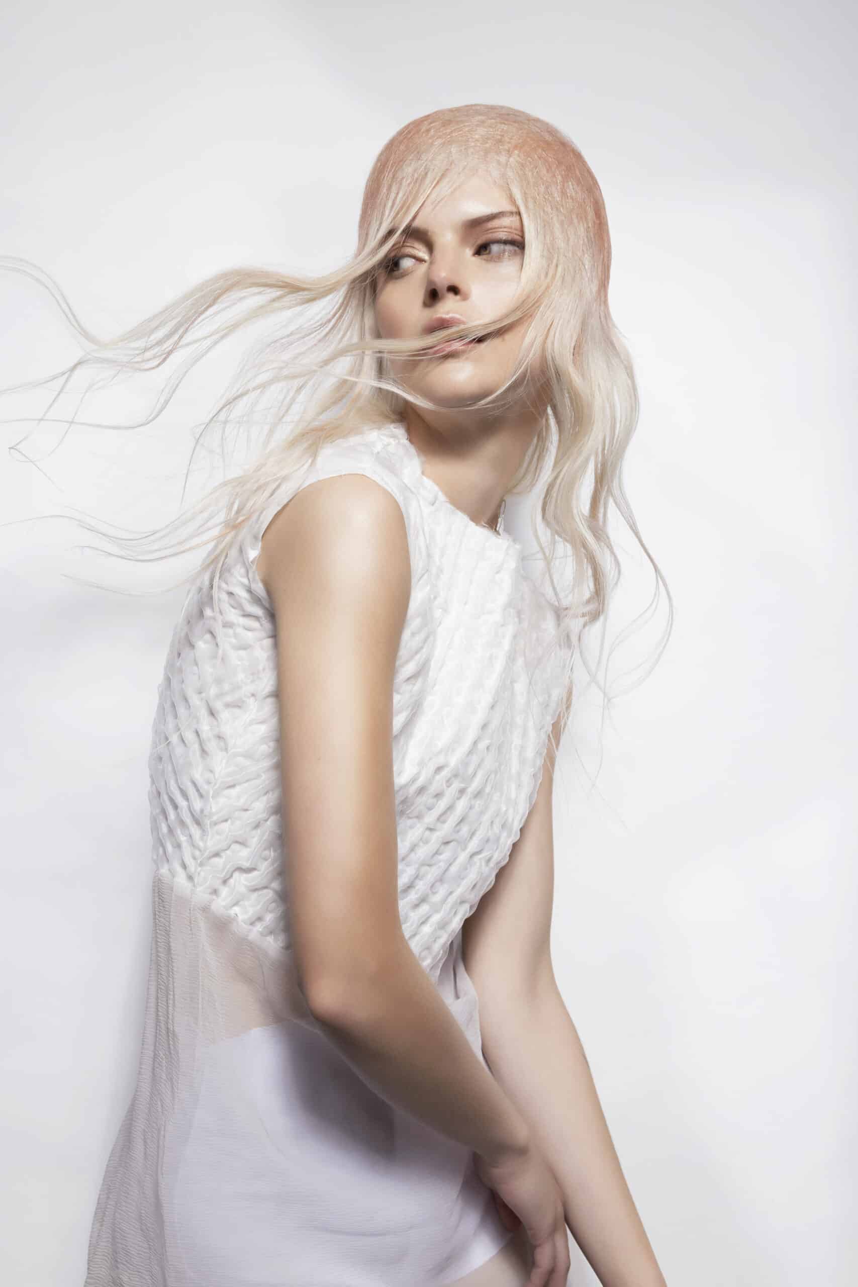 Mujer con cabello claro en movimiento y vestido blanco posando de perfil sobre fondo luminoso